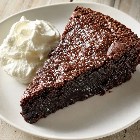En skiva kladdkaka med florsocker och vispad grädde på vit tallrik